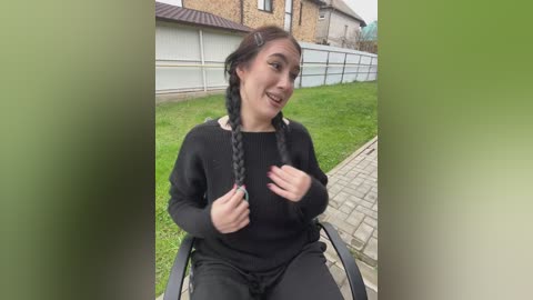 Video of a smiling, light-skinned woman with long brown hair in braids, wearing a black sweater and pants, seated on a bench in a suburban yard with a white fence and brick houses in the background.