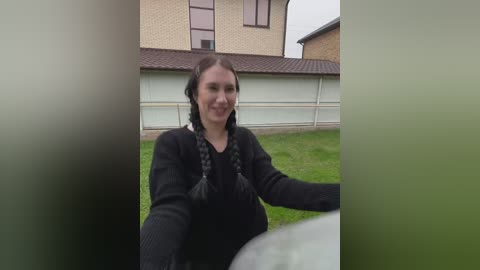 Video of a woman with long dark hair in braids, wearing a black sweater, standing on a grassy yard in front of a beige house with a garage door.