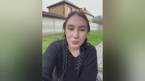 A video of a young woman with fair skin, brown hair, and wearing a black sweater, puckering her lips in a playful manner. The background features suburban houses and a grassy yard.