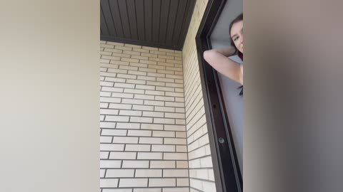Video of a person's head peeking through a glass door, with a beige brick wall and dark wooden ceiling visible. The individual has light skin, short hair, and is wearing a red shirt.