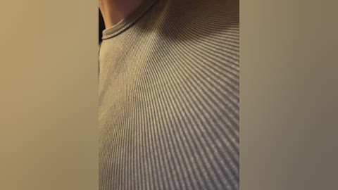 Video of a close-up view of a person wearing a grey, ribbed knit top. The background is a plain, beige wall, highlighting the fabric texture.
