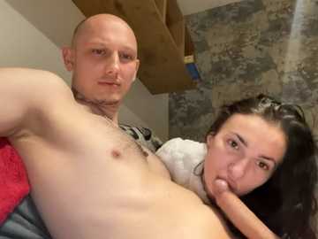 Video of a bald, light-skinned man with a toned physique, naked from the waist up, and a dark-haired woman with large, round glasses performing oral sex on him, both in a dimly lit room.