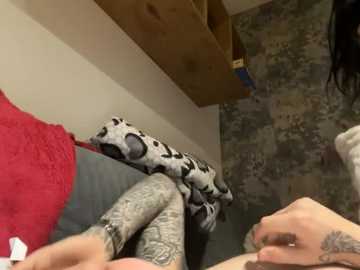 A video shows a person with long dark hair and tattoos on their arm, wearing a red top and grey leggings, sitting on a bed with a patterned pillow and wooden shelf in the background.