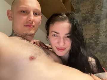 Video of a bald, muscular, light-skinned man with a tattoo on his neck, and a dark-haired, fair-skinned woman with a tattoo on her arm, both smiling in a rustic, stone-walled room.