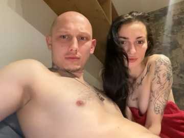 Video of a bald, muscular man and a tattooed woman with long black hair, both topless, leaning on a bed.