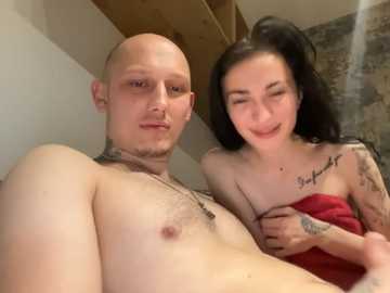Video of a topless, bald, muscular man with tattoos, holding a smiling, tattooed woman with long black hair, both wearing red towels, in a rustic room with stone walls.