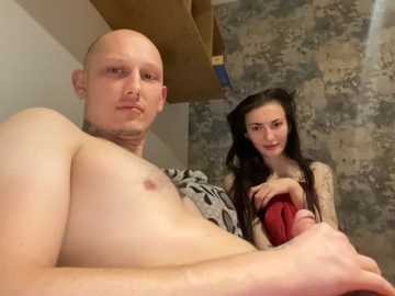 Video of a bald, shirtless man with a neutral expression, holding a red blanket, and a young woman with long dark hair, standing behind him in a rustic, stone-walled room.