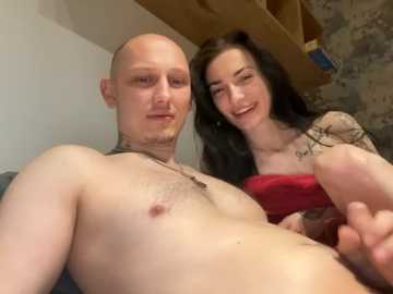 A video of a bald, shirtless man with tattoos and a smiling woman with long dark hair and a tattooed arm, both posing intimately indoors.