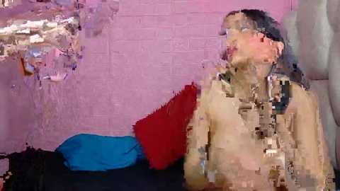 Video of a nude, light-skinned woman with long black hair, wearing glasses, sitting on a bed with a blue pillow, against a pink brick wall, looking contemplative.