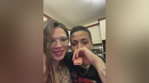 A video of two women in a kitchen. One with glasses, smiling, has long wavy hair. The other with a tattooed arm, wearing a necklace, holds a pink phone.