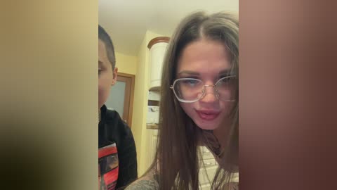 Video of a young girl with long, straight brown hair, wearing glasses, smiling at the camera in a bathroom. A boy in a striped shirt stands in the background.