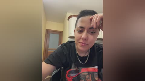 Video of a young woman with short dark hair, wearing a black graphic T-shirt and silver necklace, resting her head on her hand, in a beige-walled room with a wooden door and white lamp.