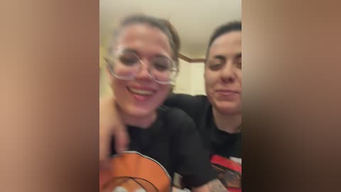 Video of two women with light skin, one with glasses, sitting closely, smiling, in a cozy setting with blurred background.