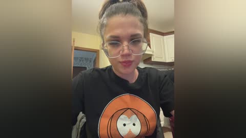 Video of a young woman with glasses, brown hair in a ponytail, wearing a black t-shirt with an orange basketball logo, indoors with white cabinets and a yellow wall.