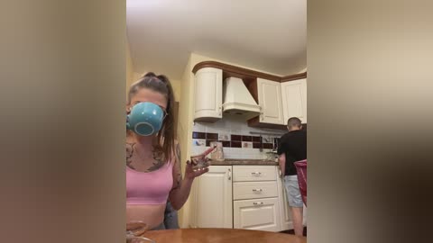 Video of a woman with a blue cup over her mouth, standing in a modern kitchen with white cabinets, brown backsplash, and a red chair.