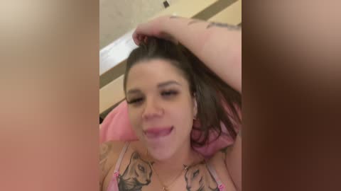 A candid video of a tattooed woman with fair skin and dark hair, wearing a pink bra, licking her finger while seated in a chair, with blurred faces in the background.