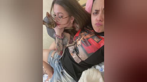 Video of a young woman with long brown hair, wearing glasses and a graphic tee, holding a brown and white cat. She's sitting on a bed with a white blanket.