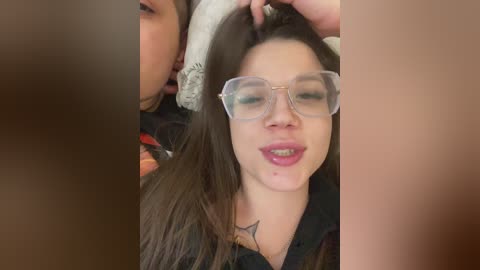 Video of a woman with fair skin, brown hair, wearing large clear glasses, smiling, with a man's hand in her hair.