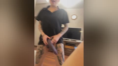 Video of a light-skinned woman with tattoos, wearing a black shirt and camo pants, holding a knife, standing in a living room with a TV and wooden floor.