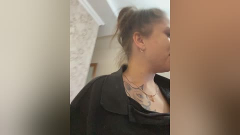 Video of a woman with a black shirt pulled down, revealing a tattoo of a bird on her chest, standing in a hallway with a marble wall and wooden door in the background.