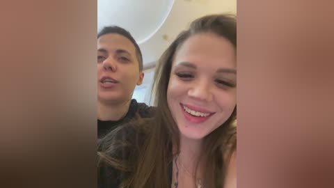 Video of a smiling, fair-skinned brunette woman with long hair, wearing a black top, and a young man with short dark hair, both with open mouths, taken indoors.