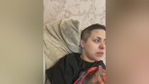 Video of a young person with short, dark hair and a buzz cut, lying on a beige quilted bedspread, wearing a black T-shirt with red and orange designs. The background features floral-patterned wallpaper.