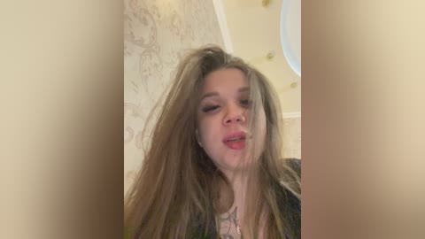 A video of a young Caucasian woman with long, messy brown hair, wearing a dark jacket, standing in a room with beige walls and a circular mirror in the background.