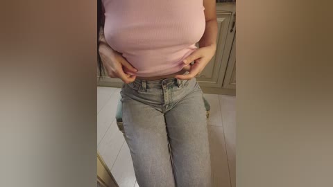 Video of a woman with fair skin, wearing a tight pink top and light-wash jeans, hands on her waist, standing in a kitchen with beige cabinets and tiled floor.