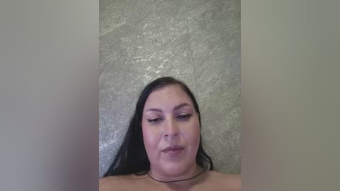 Video of a middle-aged woman with medium skin tone, dark hair, and black eyeliner, lying on a textured grey carpet, wearing a black choker necklace.