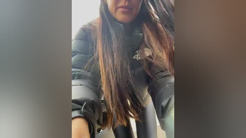 A close-up video of a woman with long, straight, dark brown hair and a light complexion, wearing a puffy, gray jacket, standing in a blurred outdoor setting.