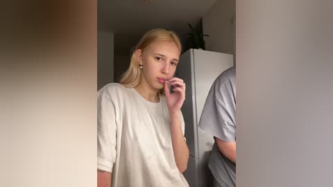 Video of a young, slim Caucasian woman with blonde hair, wearing a loose white T-shirt, standing in a dimly lit room, touching her lips, with another person partially visible in the background.
