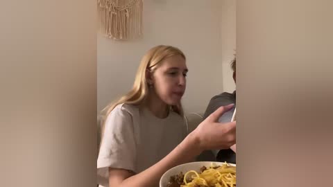Video of a blonde Caucasian woman in a beige shirt, eating noodles with chopsticks, against a cream-colored wall adorned with a macram\u00e9 hanging.