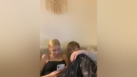 A video of two blonde women sitting on a beige couch, one wearing a black tank top, the other in a grey sweater, both holding phones, with a macrame wall hanging above.