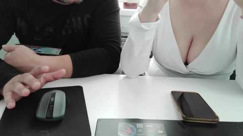 Video of a woman with large breasts in a white blouse, holding a phone, and a man with a beard and dark shirt, holding a mouse, sitting at a desk.