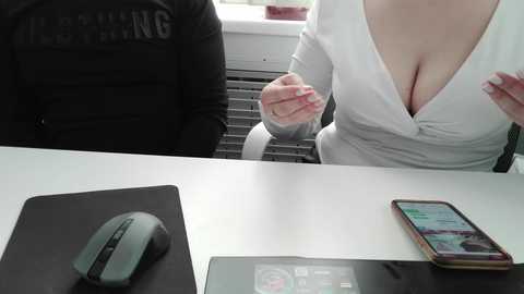 Video of a woman in a low-cut white blouse, with cleavage visible, seated at a white desk, holding a glass. A black mouse and smartphone are on the desk.
