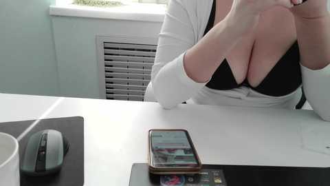 A video of a woman in a white cardigan, revealing a black bra, sitting at a white desk with a smartphone and a gray mouse.