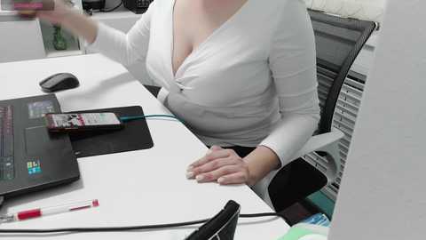 Video of a white woman in a white blouse, sitting at a cluttered desk with a laptop, phone, pen, and paper. Background shows office equipment and a black chair.