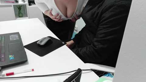 A video depicts a woman with large breasts, partially unbuttoned shirt, sitting at a desk, wearing a black jacket. A laptop and stationary items are visible on the white desk.