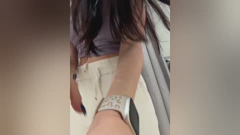 Video of a woman with long dark hair wearing a grey crop top and white high-waisted jeans, her left wrist adorned with a silver watch. Background includes a blurred white wall and a grey curtain.