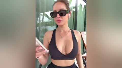 Video of a light-skinned woman with brown hair, wearing black sunglasses, a black sports bra, and black pants, holding a phone, standing in front of a glass window reflecting greenery, indoors.