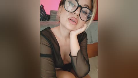 A video of a blonde woman with glasses, wearing a sheer black top, lying on a bed with a pink light in the background.