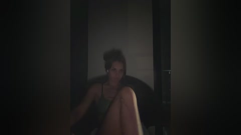 Video of a woman in a dark room, sitting on a chair, wearing a green top, with a dimly lit, shadowy background.