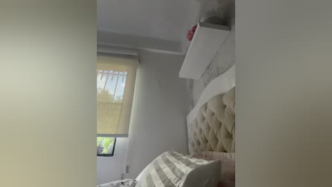 A video of a minimalist bedroom with a tufted beige headboard, white shelves, and a striped bedspread. A window with beige curtains allows natural light to filter in, revealing greenery outside.