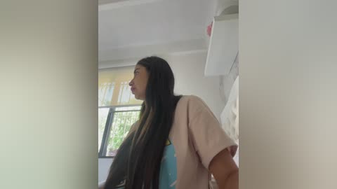 A video of a young woman with long, straight black hair, wearing a beige T-shirt, standing in a dimly lit room with a white wall and a partially visible window.