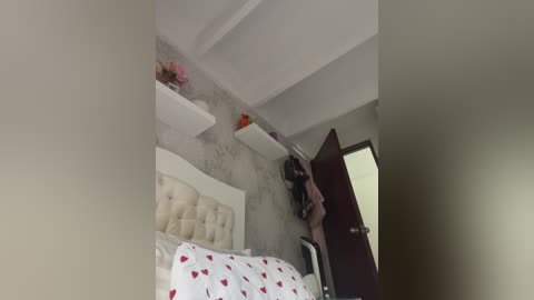 A video of a modern, minimalist bedroom featuring a white tufted headboard, white shelves, and a dark wooden door. The walls are painted light gray, and a floral-patterned duvet covers the bed.