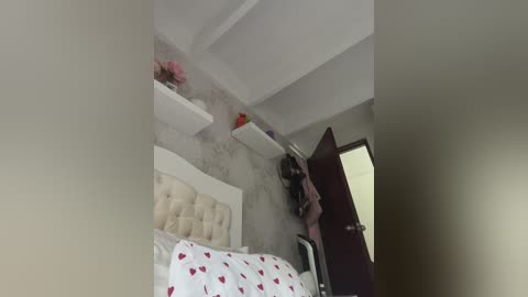 A video of a minimalist bedroom with white walls, a tufted beige headboard, and a bed with a red heart-patterned duvet. A white floating shelf holds pink flowers. A dark brown door is slightly open, revealing a dark wardrobe.