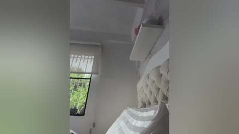 A video of a minimalist, modern bedroom with a tufted, cream-colored headboard, white shelves, and a window with sheer white curtains revealing green foliage outside. The room has a clean, neutral color palette.