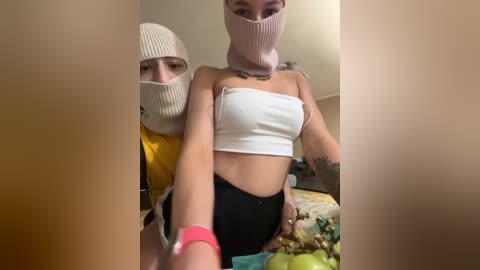 Video of two young women wearing face masks and knit hats, one in a yellow jacket, the other in a white crop top and black pants, indoors with a cluttered background.