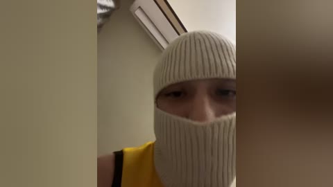 A video of a person with fair skin and short brown hair, wearing a light beige knitted mask, yellow shirt, and black wristband, standing in a dimly lit room with cream walls and a white ceiling light.