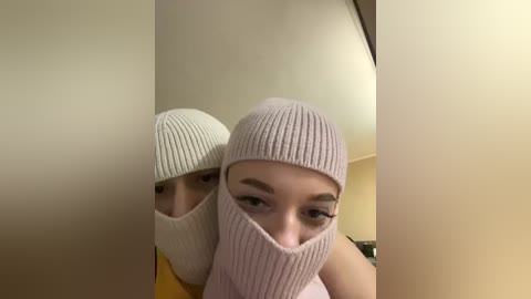 A close-up video of a young woman with fair skin and dark hair, wearing a pink knitted beanie and a matching face mask, partially obscuring her eyes. The background shows a plain beige wall and a faint reflection of her face.